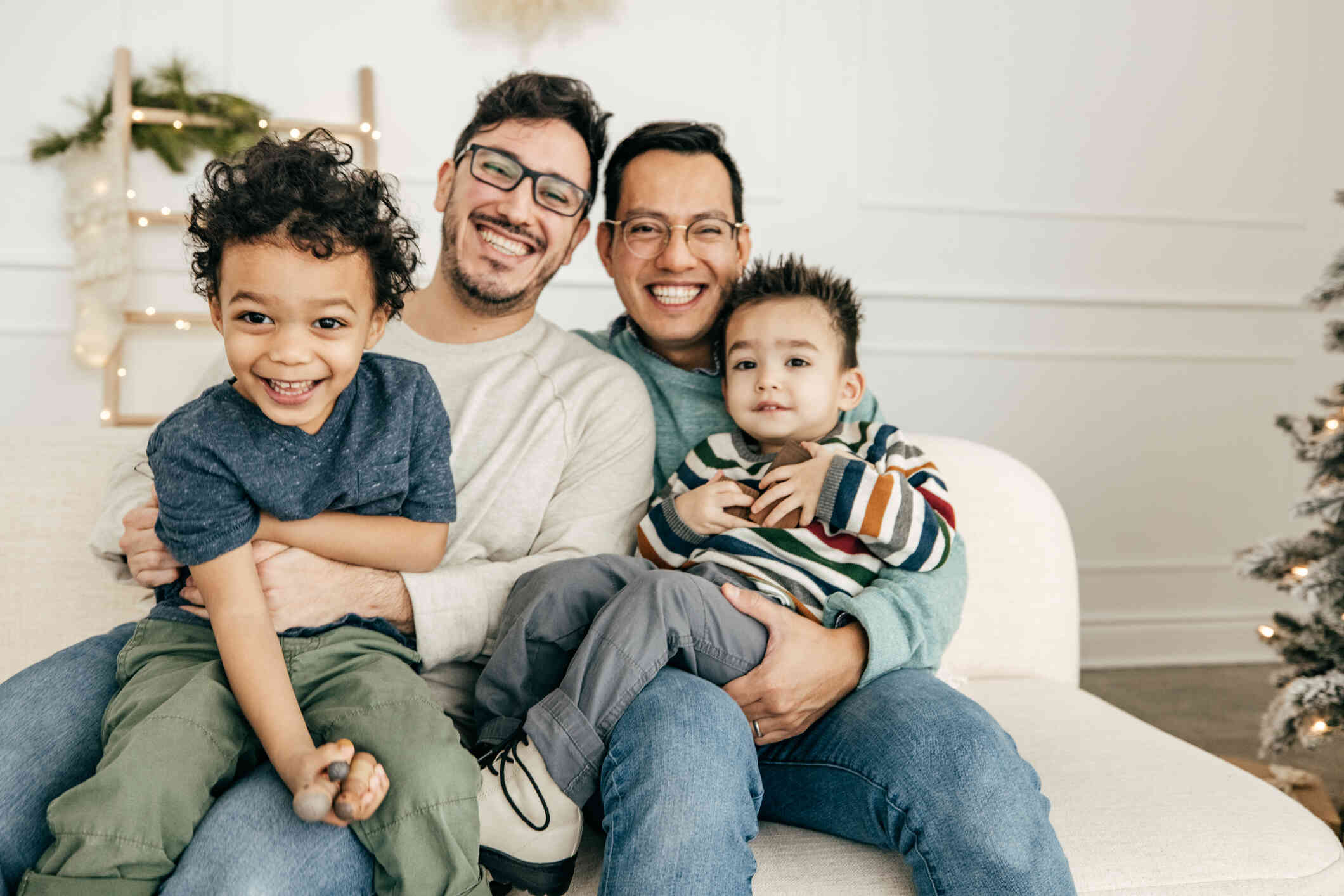 Two fathers sit in their home with their young sons in their laps as they all smile at the camera.