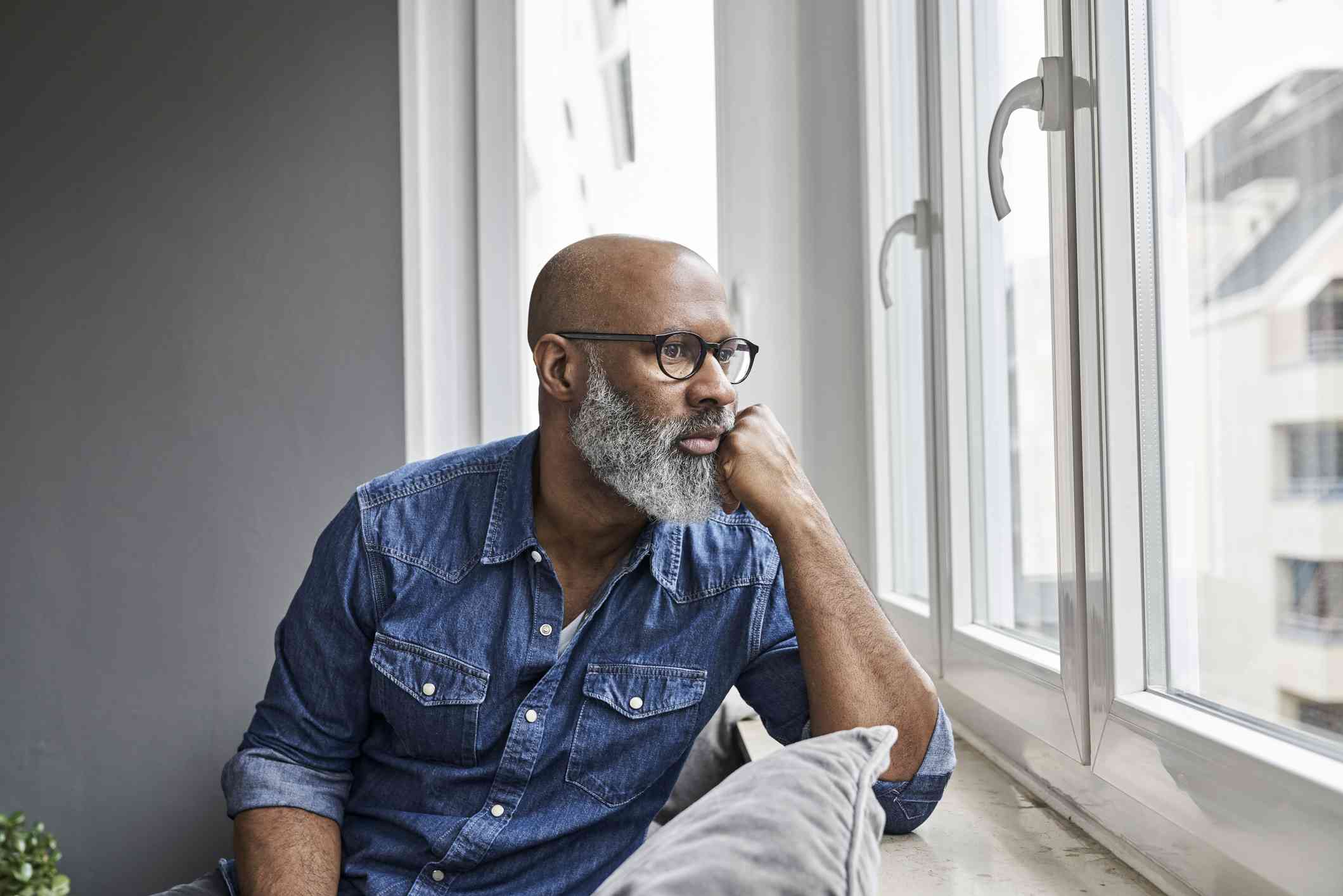 A middle aged man in a blue shirt sits near a window with his head in his hand while gazing out with a sad expression.